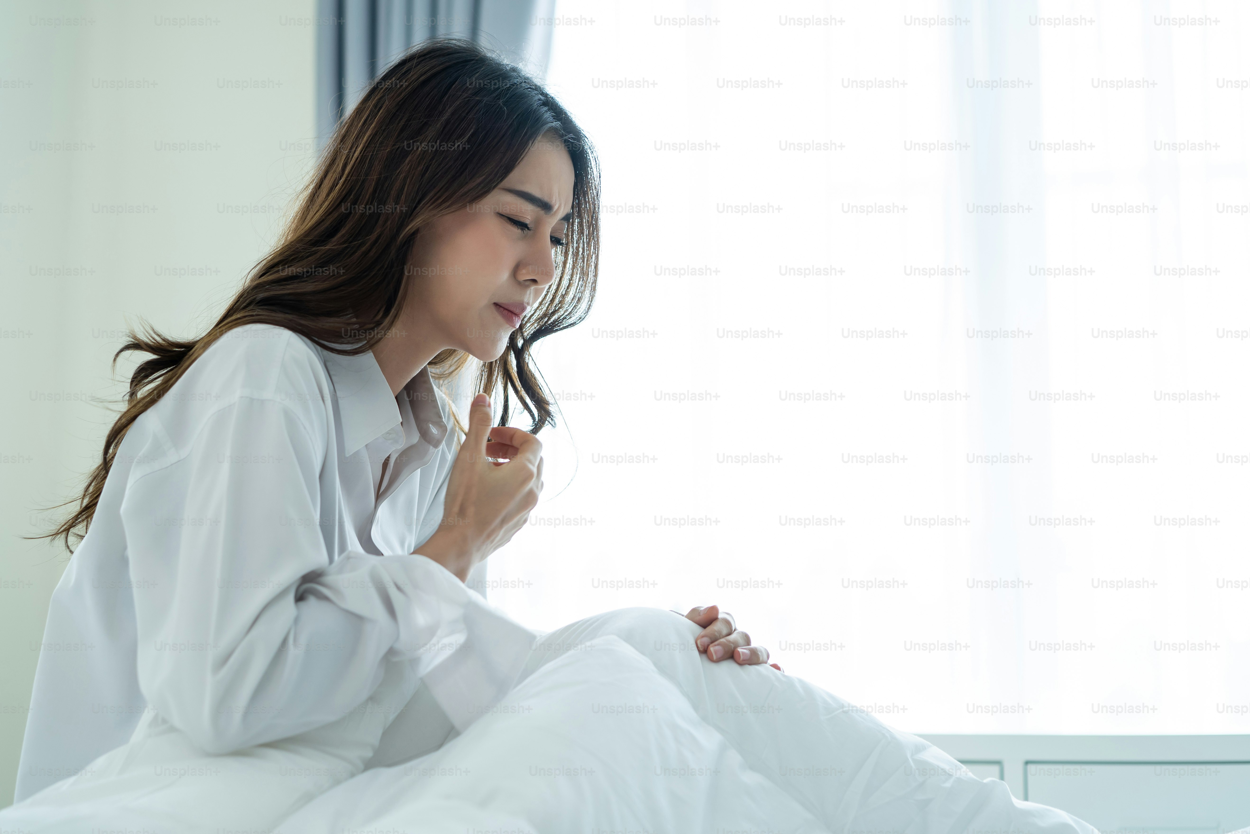 A woman in a white shirt coughing while sitting on a bed